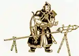 A man in black armor standing in front of a rocket, attached to a stick, with the stick being held up by two X-shaped wooden brackets.
