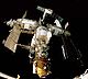 Uma estação espacial que consiste em três módulos brancos dispostos em forma de T em torno de um nó de acoplamento central. O módulo sobre o curso descendente do T tem um módulo menor, laranja ligado à extremidade distai do mesmo, e um quinto módulo projectos para trás a partir do nó de ancoragem, com um sexto módulo ligado à sua extremidade. A Progress e Soyuz são encaixadas para a estação, e cada um dos módulos projeta várias penas solares arrays, antenas e treliças. A escuridão do espaço constitui o pano de fundo para a imagem e as soleiras de um compartimento de carga do ônibus espacial pode ser visto na parte inferior da imagem.