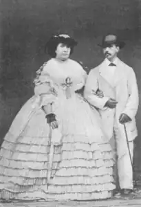 Portrait en pied en noir et blanc de la reine Isabelle II d’Espagne en robe claire et du roi consort en tunique claire avec une canne, se tenant le bras.