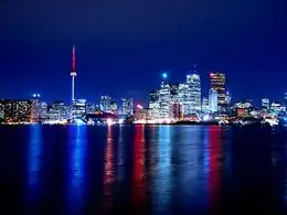 Un panorama de tours et gratte-ciels illuminés sur un ciel nocturne sans nuage et le tout se reflète dans l'eau.